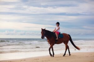A young girl wearing a safety helmet enjoys a peaceful horseback ride along the sandy shoreline of Myrtle Beach as the waves gently roll in. This scenic coastal activity highlights the unique outdoor adventures and memorable experiences available to guests staying with Casago Myrtle Beach.